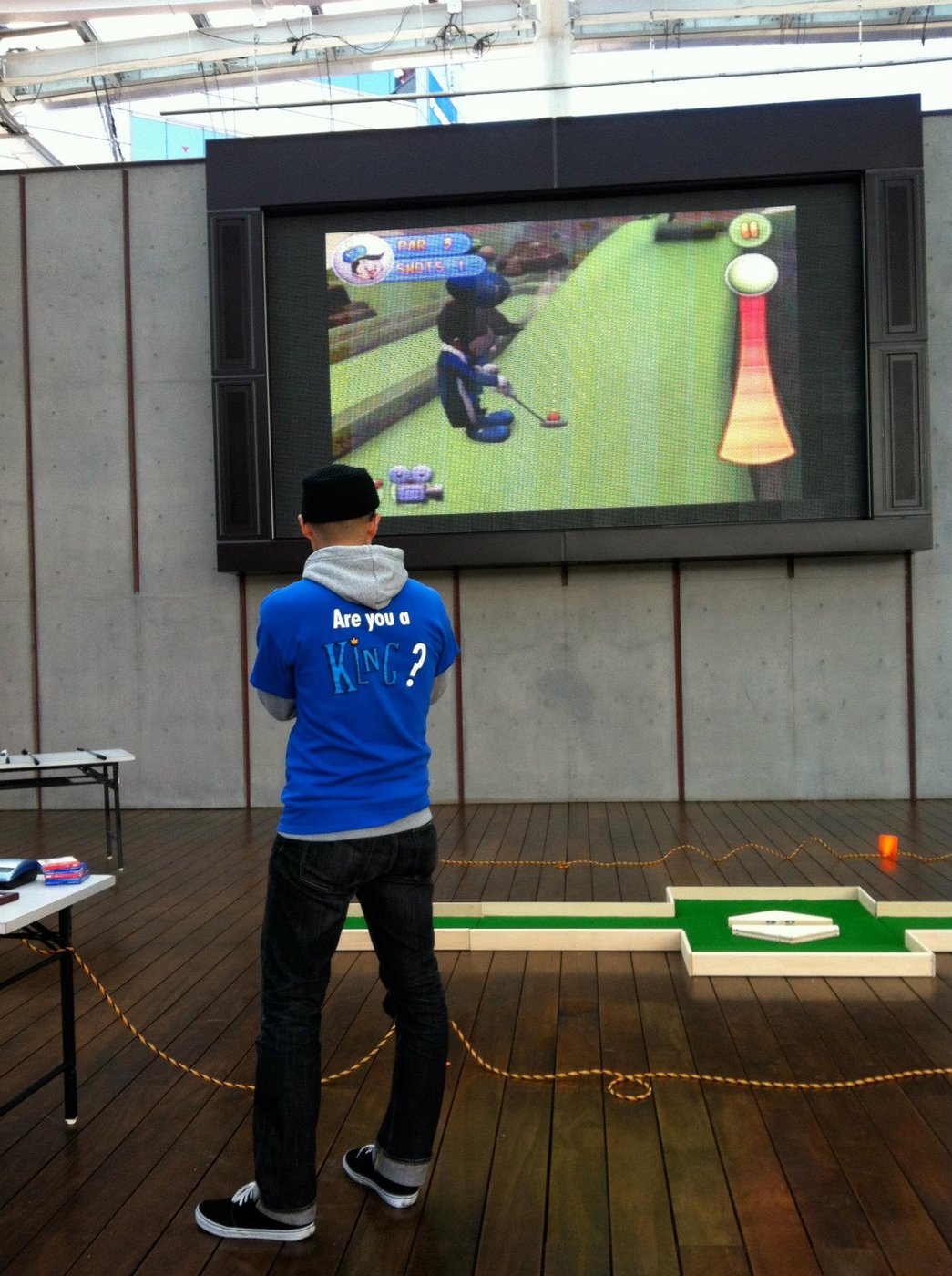 A young man in an 'Are you a KING?' t-shirt putting on a physical mini-golf hole indoors with Adventure Golf gameplay on a big screen behind him
