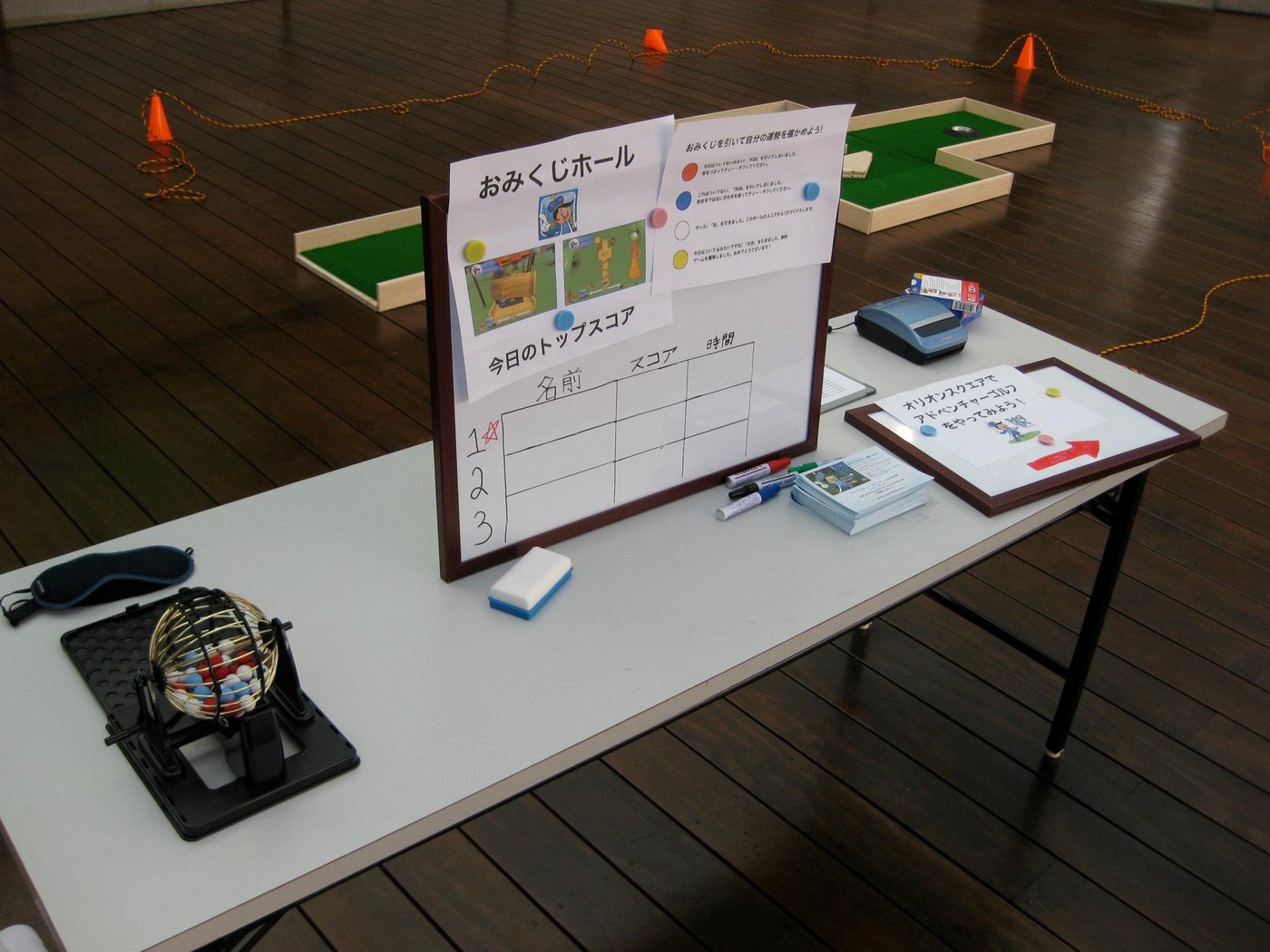A table with a printed Japanese leaderboard titled おみくじホール, a cash box, and a receipt printer, with a mini-golf hole visible behind