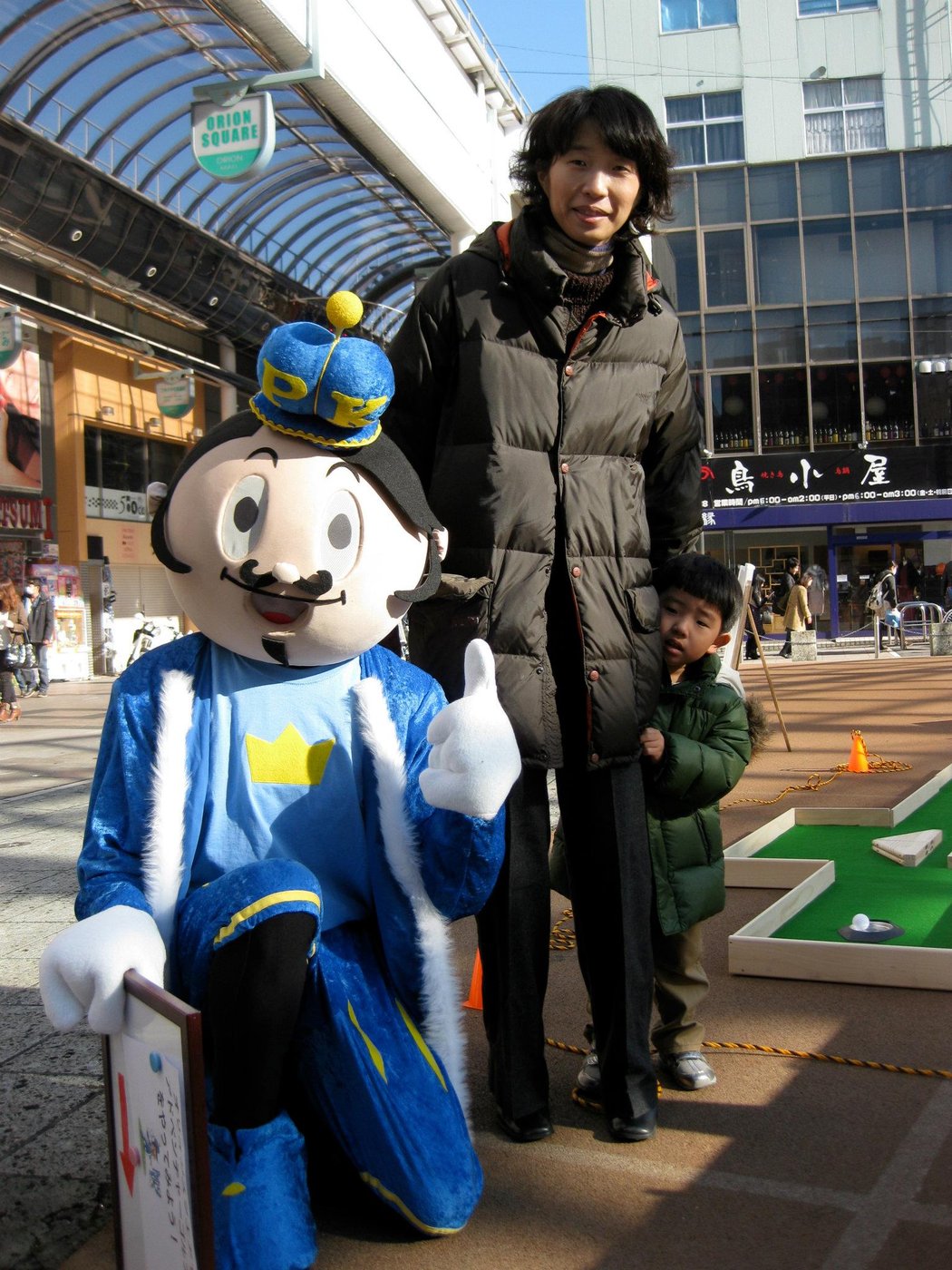 A Japanese mother and young child posing with Putty the mascot in a shopping arcade, with a pop-up miniature-golf hole visible behind