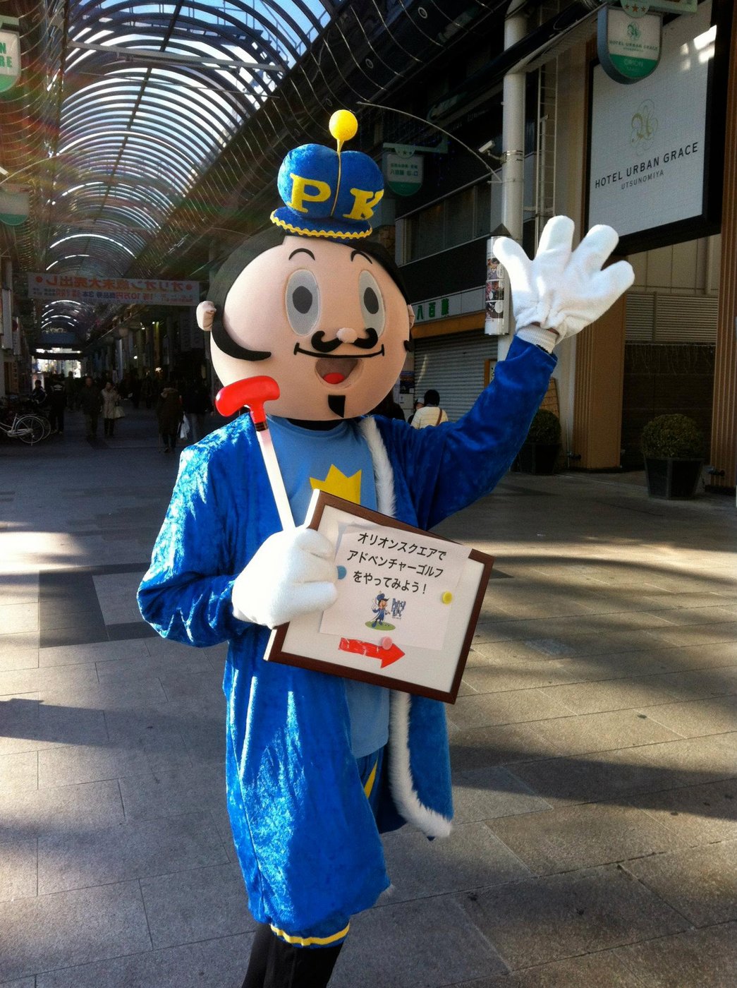 Life-size Putty mascot in a Japanese shopping arcade, waving and holding a Japanese-language sign