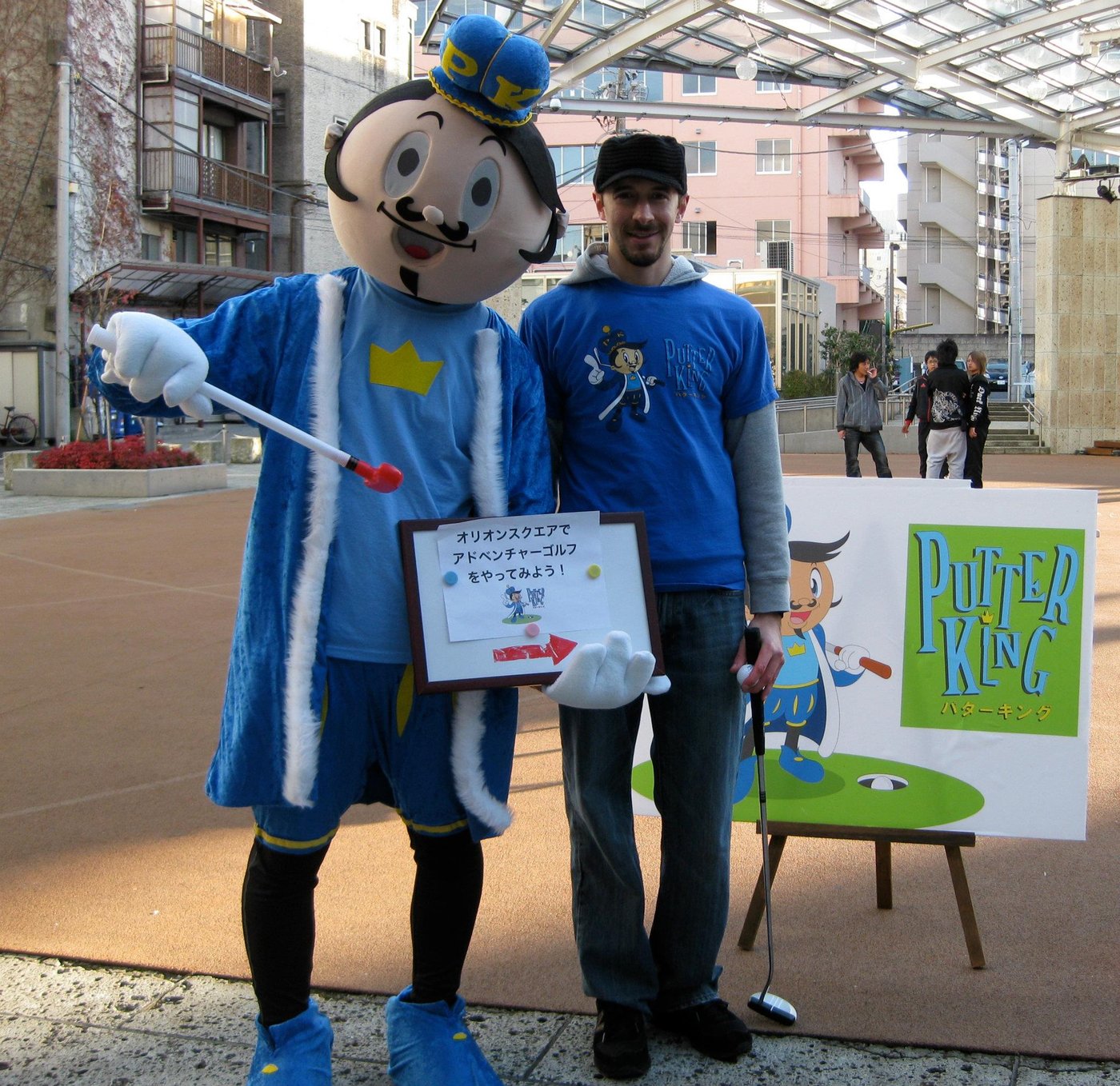 The Putty mascot standing with a team member in a Japanese plaza in front of a Putter King easel sign