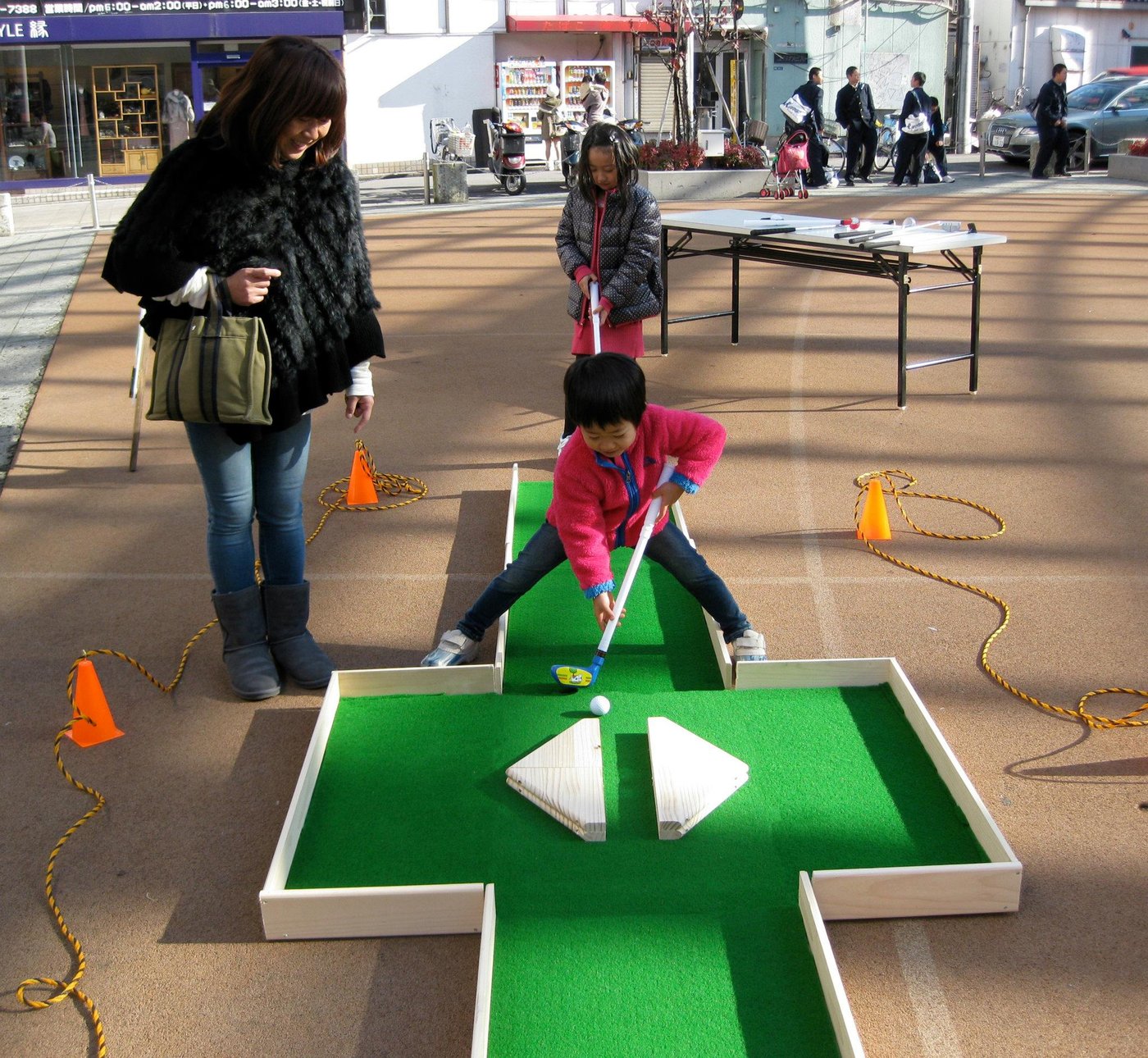 A young girl in a pink coat mid-swing on the pop-up mini-golf hole, with adults watching
